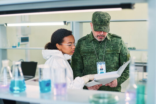 Young woman in hazmat suit explaining something in secret document to mature military man looking through description of experiment