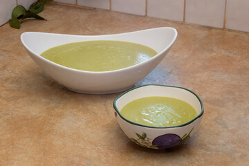 A rustic kitchen counter with a tiled surface showcases an elegant oval dish, featuring a deep bowl adorned with delicate floral patterns, both brimming with creamy broccoli soup.