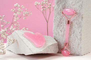 Rose quartz gua sha massage scraper and face massage roller on a natural stone plinth. Facial care