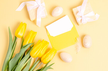Easter eggs, gift boxes, an empty postcard in an envelope and tulips on a yellow background.