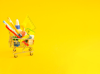 Stationery accessories in a shopping trolley on a yellow background with place for text.