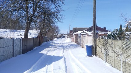 After the snowfall, there is snow on the road, trees and roofs of houses. Cars have made two ruts in the snow. Along the street there are concrete, brick and wooden fences, trees and a power line 
