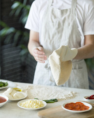 Process of making pizza. Pizzaiollo in white apron kneads and shapes dough. Ingredients for pizza: cheese, sauce, pepper, flour on table. Pizza base. Pizzeria kitchen. Front view. Soft focus. 