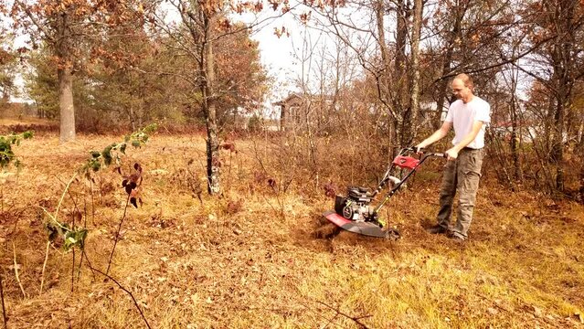 Man in white shirt using gas tiller to cultivate fresh garden bed in his woody back yard.