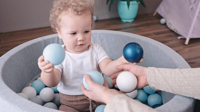 Happy mother playing with her baby in soft ball pit pool with colorful balls, caucasian baby boy having fun in kids dry pool for fun games. Slow motion