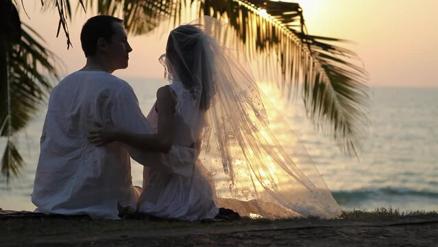 Newlyweds enjoy atmosphere of sunset against sun-path on ocean surface. Bride strokes groom under palm branch on beach after ceremony