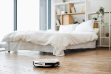 Obraz na płótnie Canvas Efficient robotic vacuum cleaner removing dust from hard flooring against cozy sleeping room background . Economical wireless device keeping large spaces at home constantly tidy during days.