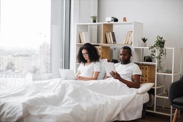 African american romantic partners using electronic devices while lounging on wide bed near panoramic window. Beautiful woman with laptop and relaxed man with tablet enjoying leisure time at home.