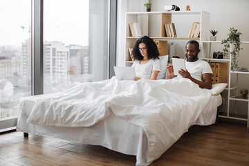 African american romantic partners using electronic devices while lounging on wide bed near panoramic window. Beautiful woman with laptop and relaxed man with tablet enjoying leisure time at home.