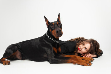 A young beautiful Doberman with a chain around his neck and his owner, elegant girl in a long black coat and bright red lipstick. Isolated on a white background.