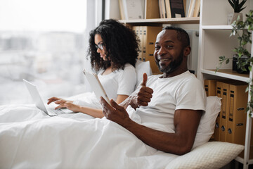 Smiling multiethnic male scrolling through webpages on tablet in bed while busy lady typing on portable computer on background. Mature people in relationships spending quality time together at home.