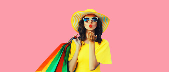 Portrait of beautiful happy young woman with shopping bags blowing her lips sending sweet air kiss wearing summer straw yellow hat on pink background
