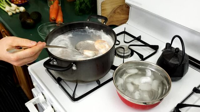 Medium size shrimps being stirred while cooked in pot of hot water over gas stove while bowl of ice water prepared for cooling down sea food.