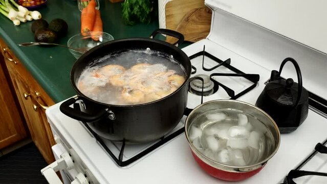 Medium size shrimps being cooked in pot of hot water over gas stove while bowl of ice water prepared for cooling down sea food.