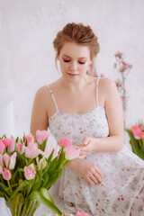 A beautiful woman feels sitting among the flowers in a white studio, gently touches the petals of a tulip with her hand.
