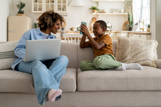 Happy African American Family Mother And Son Enjoy Weekend At Home Rest On Sofa With Gadgets. Little Boy Child Showing Something On Smartphone To Busy Mom Freelancer, Distracting Her From Remote Job