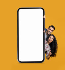 Mobile Mockup. Cheerful Arab Couple Peeking Out Behind Big Blank Smartphone