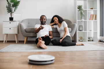 Positive african man and woman sitting together on floor and using remote controller for robot...