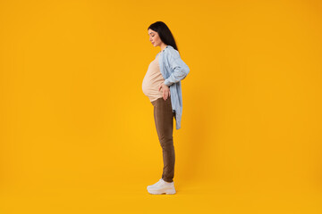 Fototapeta na wymiar Full body length shot of young pregnant woman standing over yellow studio background, side view, free space