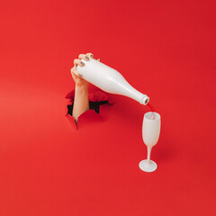 A female hand comes out from the red paper and holds white bottle with champagne glasses.