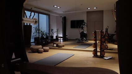 rear tracking shot of a gym and fitness room with a large mirror as a wall, a mat on the floor, weights, a machine in the background, a parquet floor and dimmed lighting, decorative potted plants