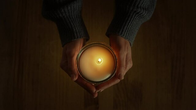 Female hands holding candle appear in the frame, over a wooden table background. Moment of sorrow and grief. Spiritual significance of holy sacrament brings peace and meditation, inspiring to pray.