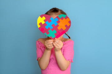A shy timid girl in a pink T-shirt covers her face with a heart made of colorful puzzles. To tell ,...