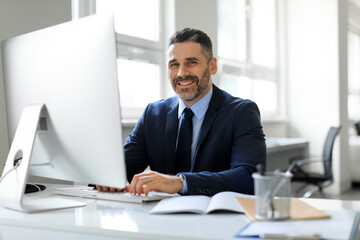 Happy middle aged businessman in suit sitting in office and smiling at camera, working with modern computer, free space