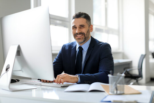 Happy middle aged businessman in suit sitting in office and smiling at camera, working with modern computer, free space