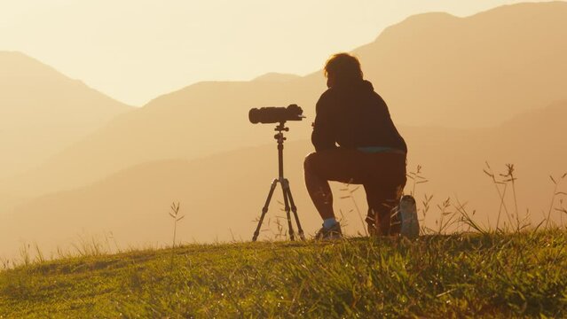Photographer on the hill. Young male photographer takes pictures with camera and telephoto lens set on tripod on the green hill in the mountains at sunrise
