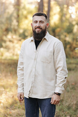 Portrait of young smiling bearded man in white casual jacket looks at camera on autumn nature background in countryside or in park. . Concept of style, walking in fresh air and unity with nature.