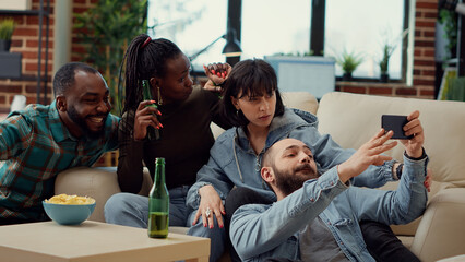 Group of friends taking picture on mobile phone app, having fun with photos at house party with beer bottles. Taking selfies and making memories with internet smartphone. Handheld shot.