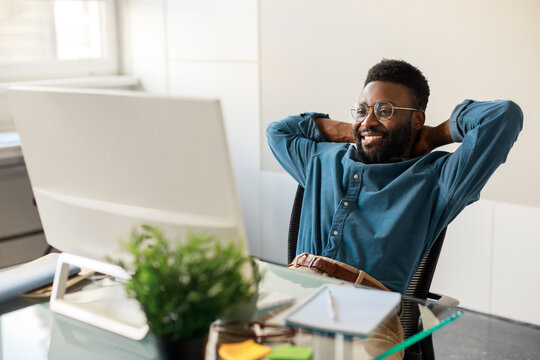 Happy black businessman taking break and enjoying result of project, sitting at table, looking at computer screen