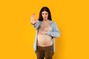 Young angry pregnant woman touching belly and doing stop sing with palm of hand, yellow background