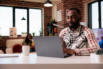 African american programmer writing code typing on laptop keyboard while girlfriend is reading a book in home living room. Male freelancer working remote focused on portable computer screen.