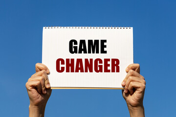 Game changer text on notebook paper held by 2 hands with isolated blue sky background. This message can be used as business concept about game changer.