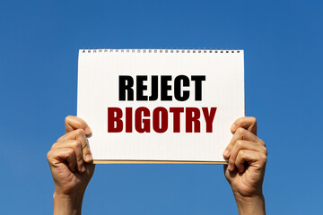 Reject bigotry text on notebook paper held by 2 hands with isolated blue sky background. This message can be used as business concept about reject bigotry.