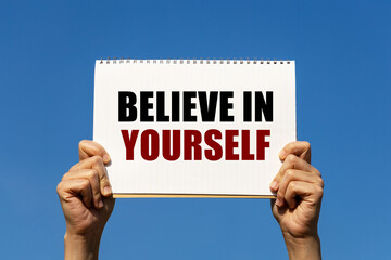 Believe in yourself text on notebook paper held by 2 hands with isolated blue sky background. This message can be used as business concept about believe in yourself.