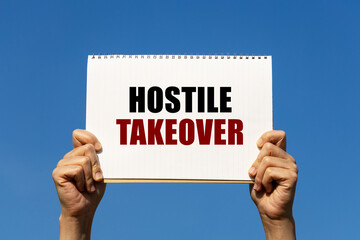 Hostile takeover text on notebook paper held by 2 hands with isolated blue sky background. This...