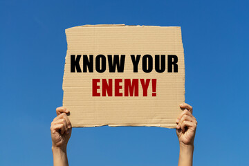 Know your enemy text on box paper held by 2 hands with isolated blue sky background. This message...