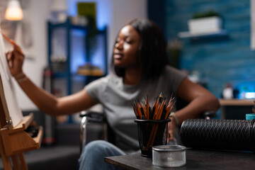 Close up of jar of colored pencils on desk in plastic arts workshop. African american professional sketcher living with disability in legs making drawing on canvas easel at artistic studio.
