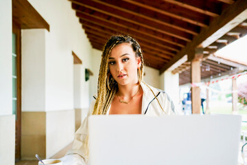 Female entrepreneur with stylish braids sitting at table with laptop and working remotely in cafe while looking at camera