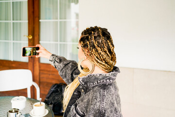 A woman taking a picture with her phone with braids in her hair and a ring in her ear, wearing a jeans jacket, in the background a wooden door with glass