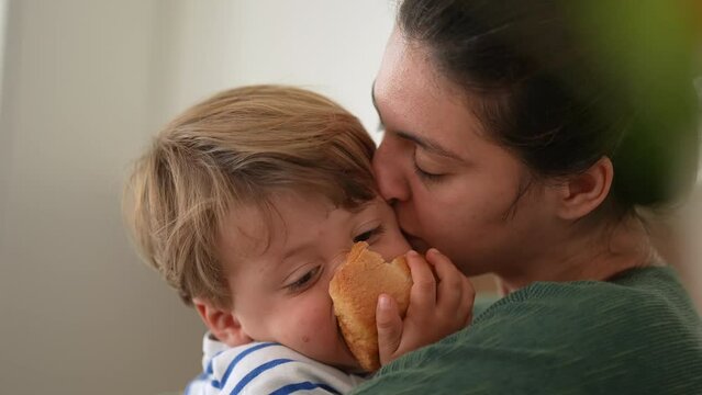 Affectionate mother kissing little boy son in cheek while eating bread. Parenting family happiness concept. Mom embracing and hugging child. Candid lifestyle