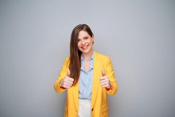 Smiling woman in yellow jacket on gray background showing thumbs up.