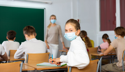 Teenage girl in face mask turned around and looking at camera during lesson in classroom.