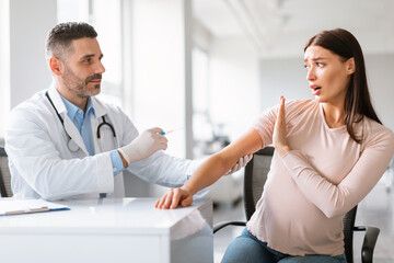 Scared pregnant lady refusing vaccine injection, sitting at clinic and showing stop gesture