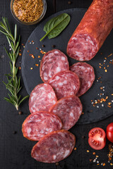 Salchichon sausage sliced on a stone board with spices, cherry tomatoes and mustard on a black wooden table.