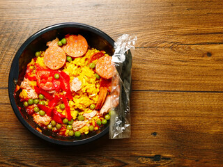 Ready made Spanish paella in a black plastic dish for microwave on a wooden table. Premium quality meal for take away or on the go. Food with rice and shrimp and chorizo.
