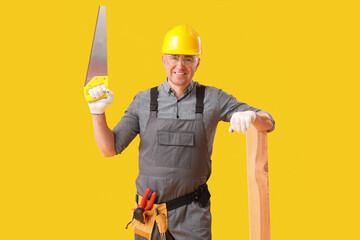 Mature carpenter with hacksaw and wooden plank on yellow background